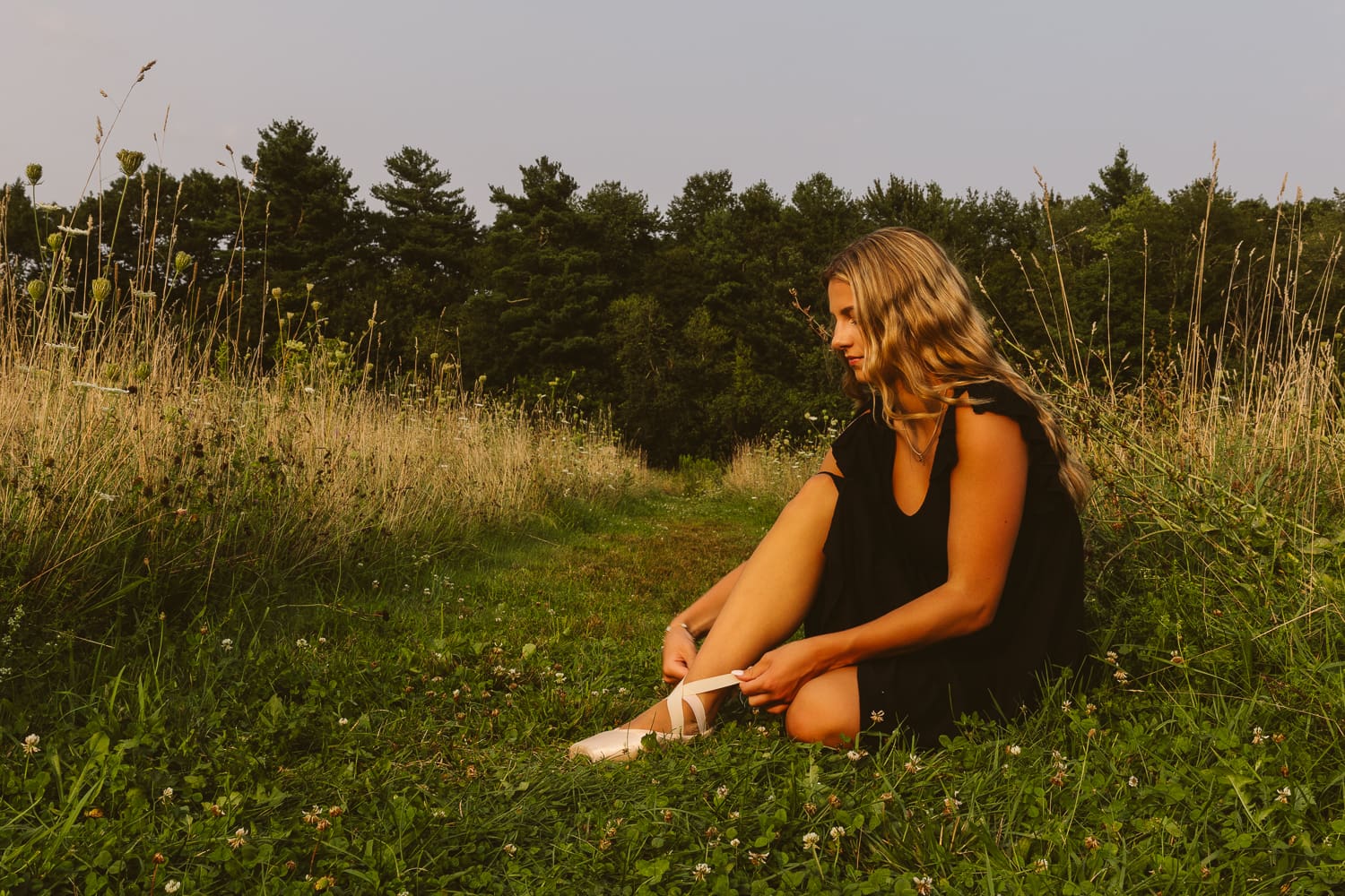 High school senior girl sitting in a grassy field at golden hour, adjusting ballet pointe shoes during an editorial senior portrait session in Massachusetts with Jennifer Helmka, Photographer.