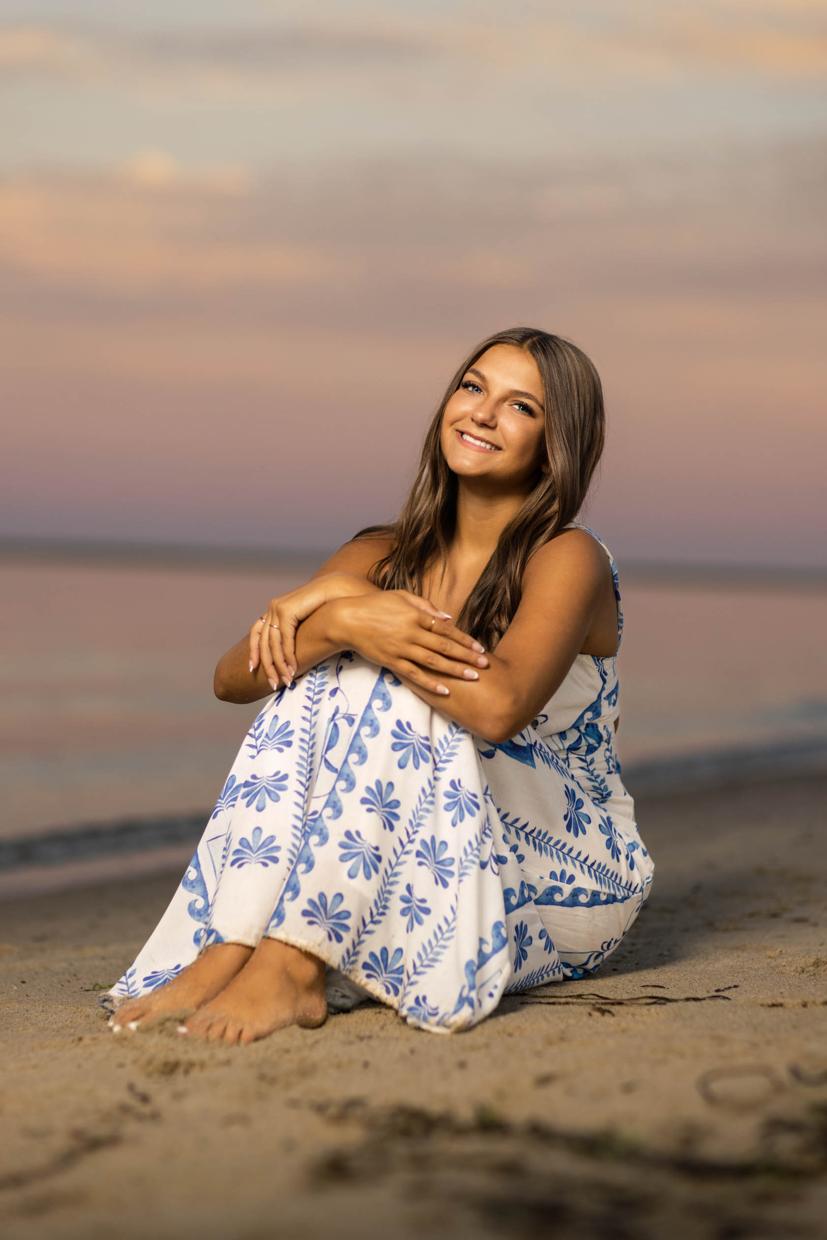 Editorial senior portrait of high school senior wearing a white maxi dress with blue designs sitting on the beach during blue hour during a senior photography session in Chatham, Cape Cod, Massachusetts with Jennifer Helmka, Photographer.