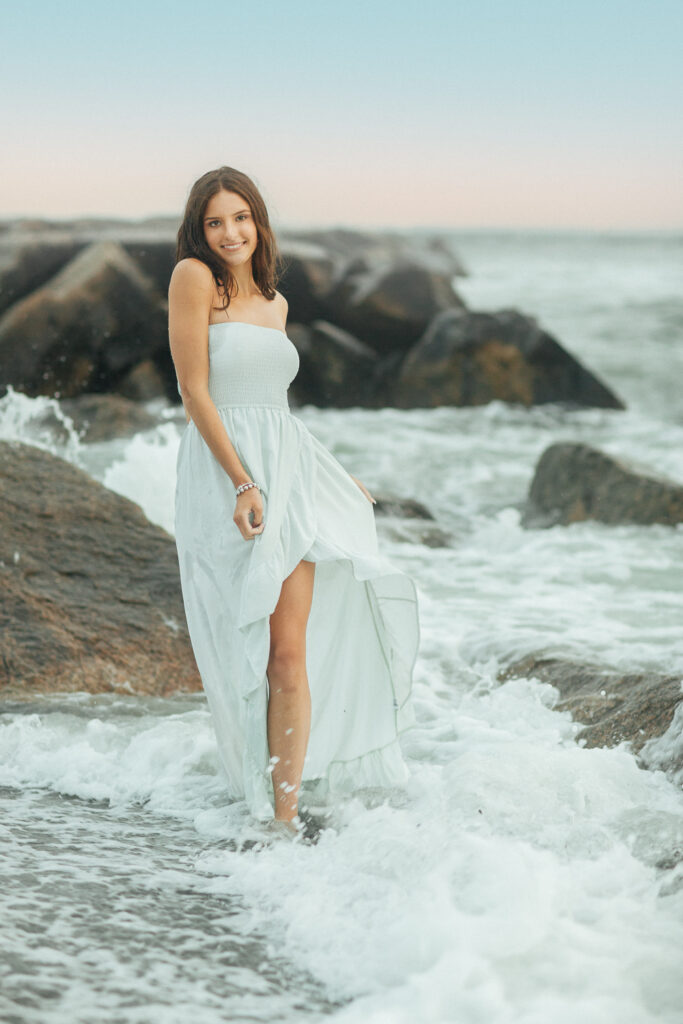 Editorial senior portrait of a high school senior wearing a light blue flowing maxi dress standing among ocean waves during a beach session at Brenton Point in Newport Rhode Island with Jennifer Helmka, Photographer.