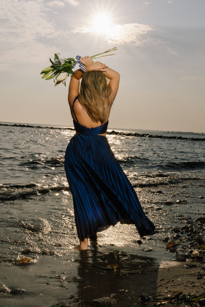 Editorial senior portrait of a high school senior wearing a navy pleated maxi dress standing barefoot at the shoreline during a coastal senior session with Jennifer Helmka, Photographer.