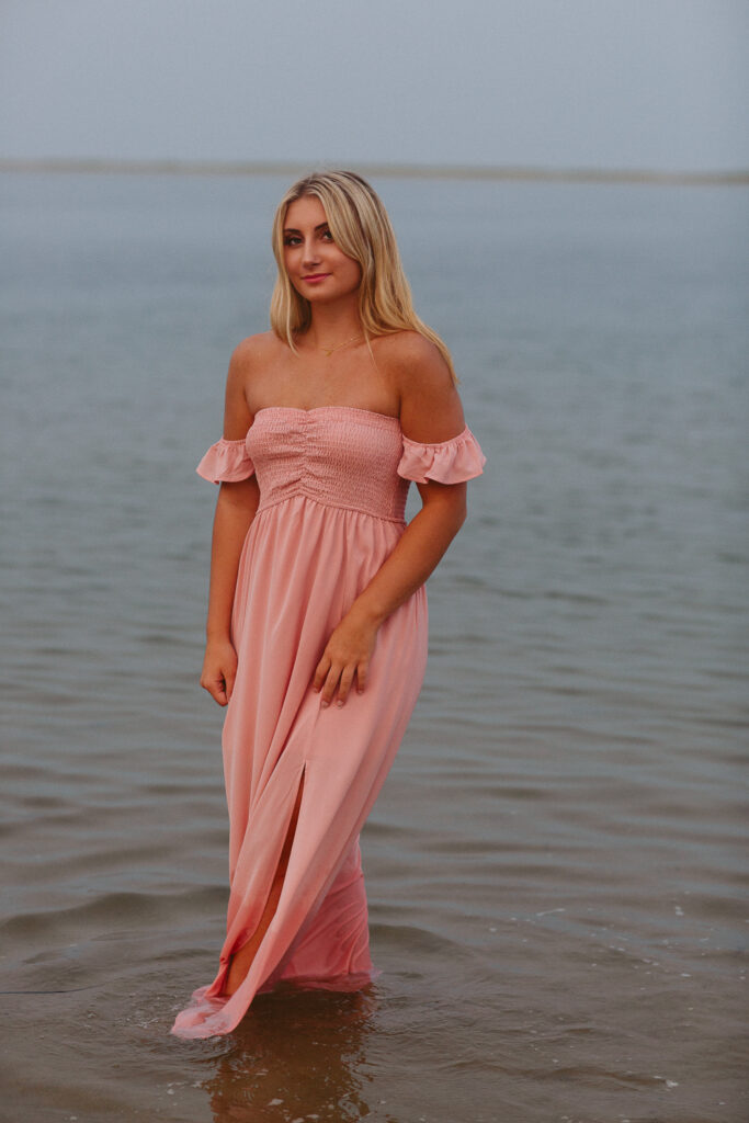 Editorial senior portrait of a high school senior standing in calm ocean water wearing a blush pink maxi dress during sunset in Chatham, Cape Cod, Massachusetts with Jennifer Helmka, Photographer.