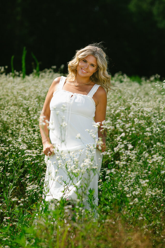 Editorial senior portrait of a high school senior wearing a flowing white maxi dress surrounded by wildflowers during a senior photography session in Massachusetts with Jennifer Helmka, Photographer.